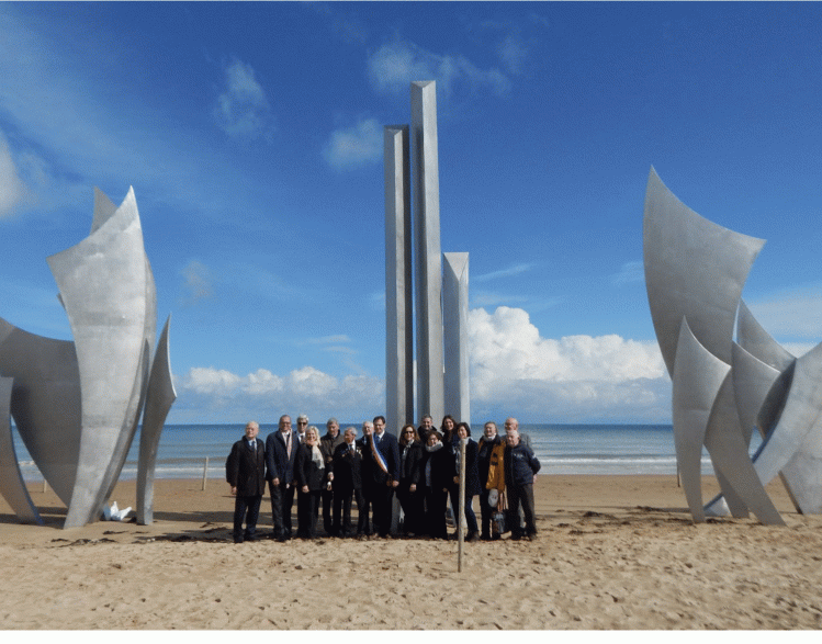 Delegation from Omaha Sister Cities and local leaders stand on Omaha Beach in 2019 in front of the monument, Les Braves
