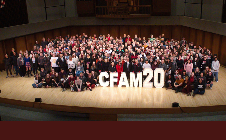 Students, staff, and faculty from the College of Communication, Fine Arts, and Media on a stage surrounding a big sign that says