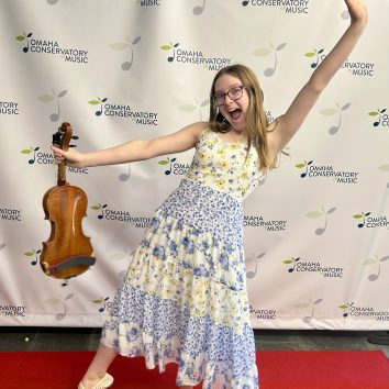 Classical Kid, Sara, poses in front of an Omaha Conservatory of Music Backdrop. She has a huge smile and holds her violin and has her other hand in the air.